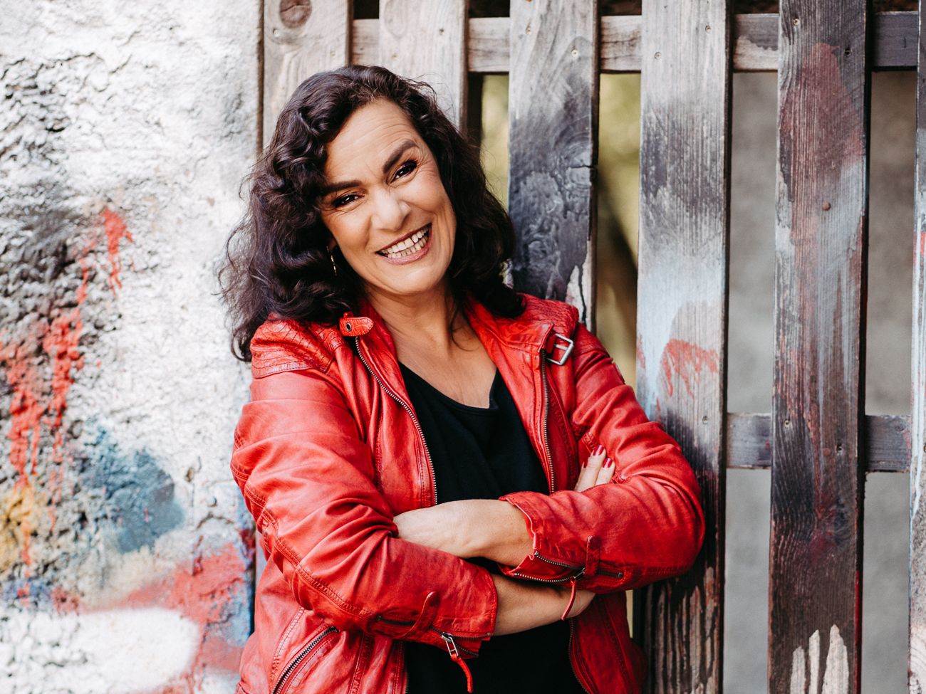 Katharina Maehrlein lachend in roter Lederjacke mit verschränkten Armen vor einer Graffiti-Wand